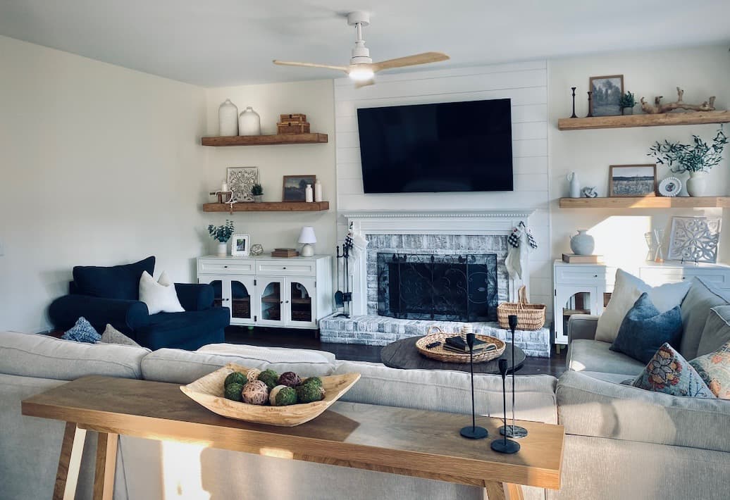 Living room with a wall-mounted TV above a whitewashed brick fireplace, floating shelves, and a navy accent chair