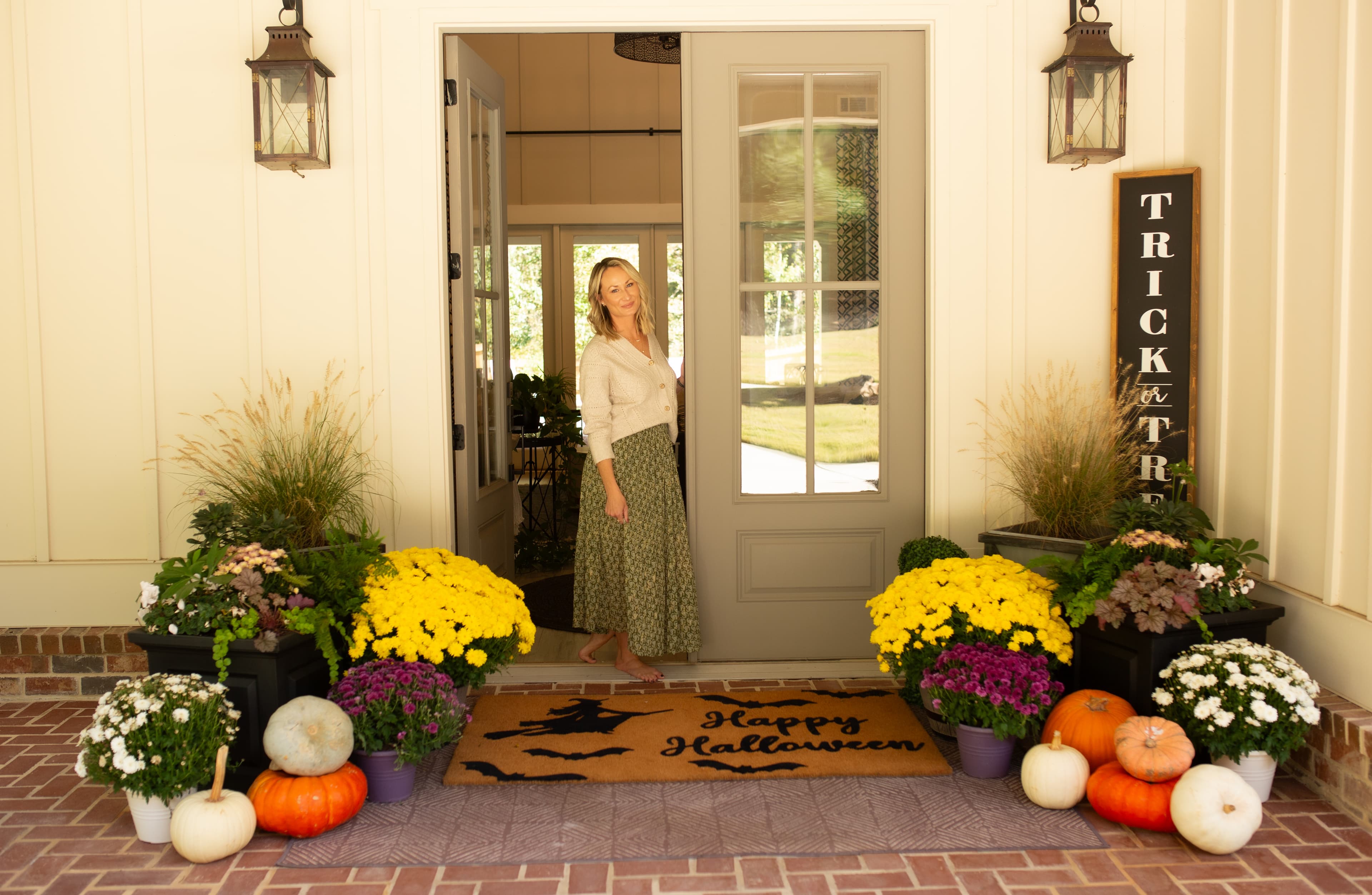 Lindsey Young standing in a front doorway styled for Halloween with mums, pumpkins, and a Happy Halloween doormat